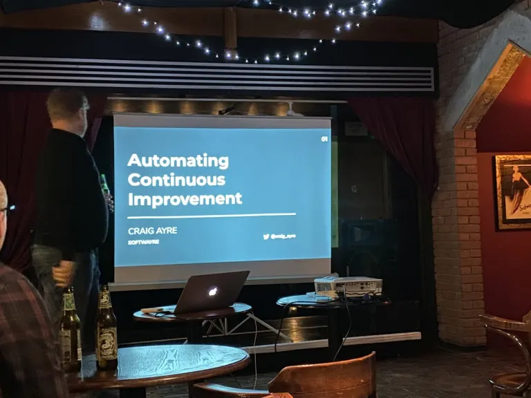 Craig stands beside a projector screen showing the opening slide for Automating Continuous Improvement, with a laptop and projector set up in front of him at a pub venue.